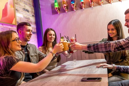 Group of 5 young people, 3 ladies and 2 men touching glasses across the table to say cheers.